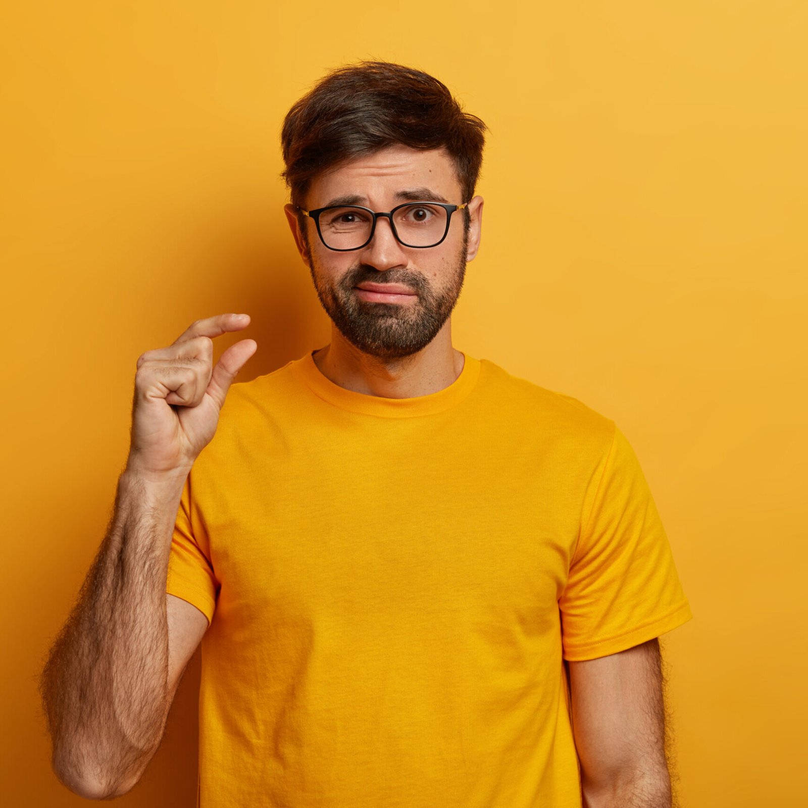 Bearded man makes hand gesture, advice to have small amount of something, purses lips, suggests spend some time together, dressed in yellow t shirt and eyewear, poses indoor. Size and measuring