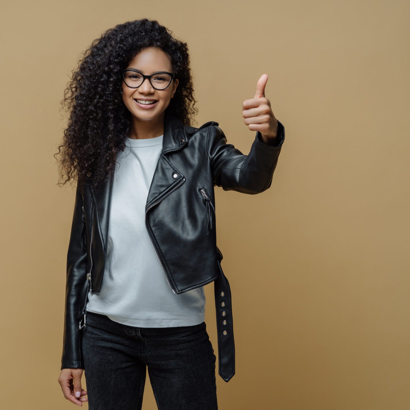 Confident cheerful African American woman shows thumb up in approval, gives recommendation or advice dressed in stylish leather jacket smiles positively isolated on beige background. I like and accept